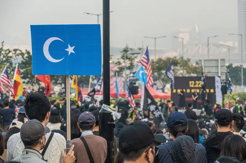 Rectangular blue East Turkestan crescent-and-star sign on a wooden stick, held above a crowd seen from behind.
