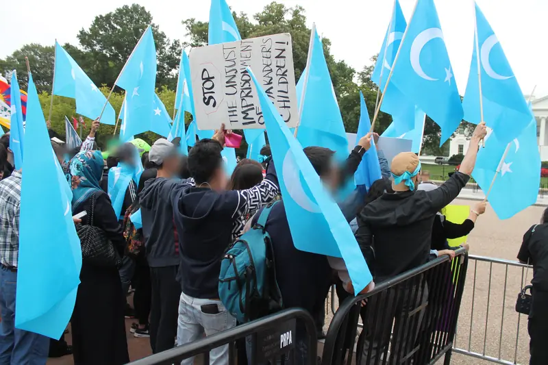 Crowd behind barriers holding dozens of pale blue East Turkestan flags and a hand-lettered placard.